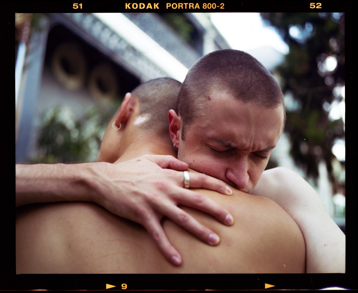 A man with closed eyes embraces another man, resting his head on his shoulder.