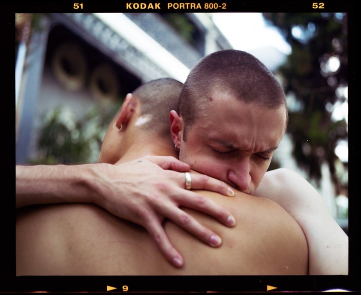 A man with closed eyes embraces another man, resting his head on his shoulder.