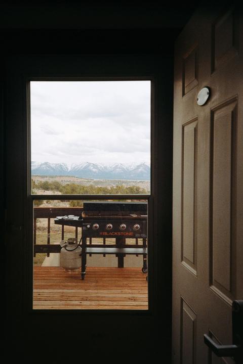 A black griddle and a propane tank sit on a wooden deck, with snow-capped mountains visible in the distance through a window.