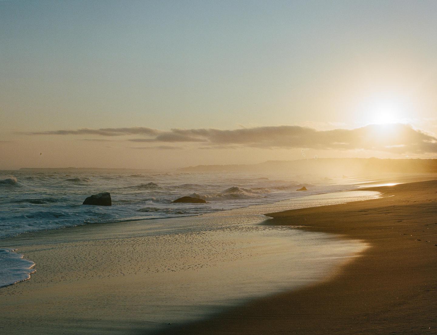 The sun sets over a beach with waves crashing on the shore and large rocks in the water.