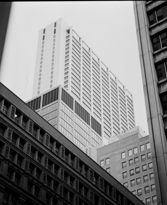 A tall, modern building with many windows rises above other buildings, framed by a dark building on the left and a lighter building on the right.