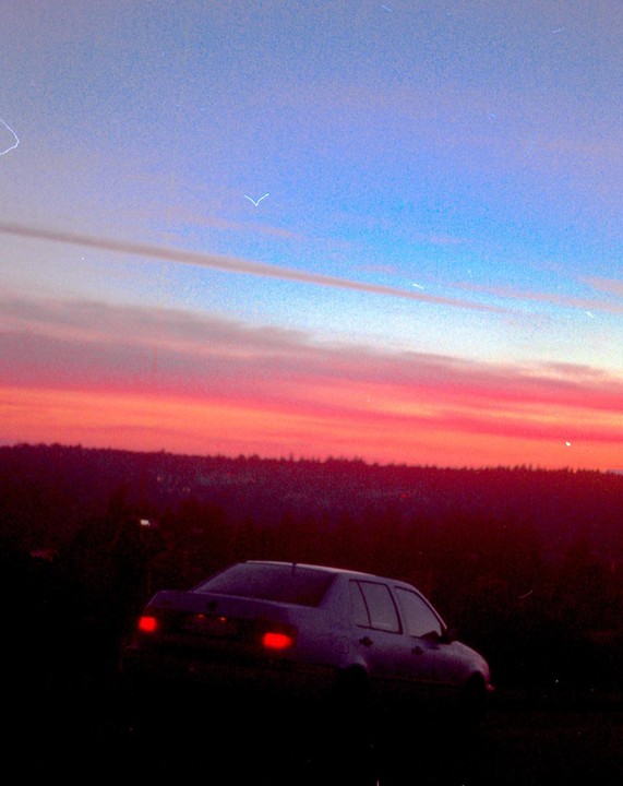 A car is parked on a hill overlooking a forest at sunset.