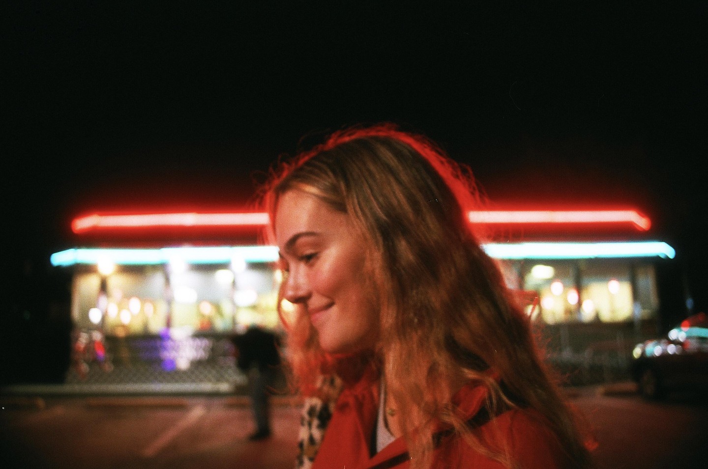 A woman with long hair smiles while standing in front of a building with neon lights at night.