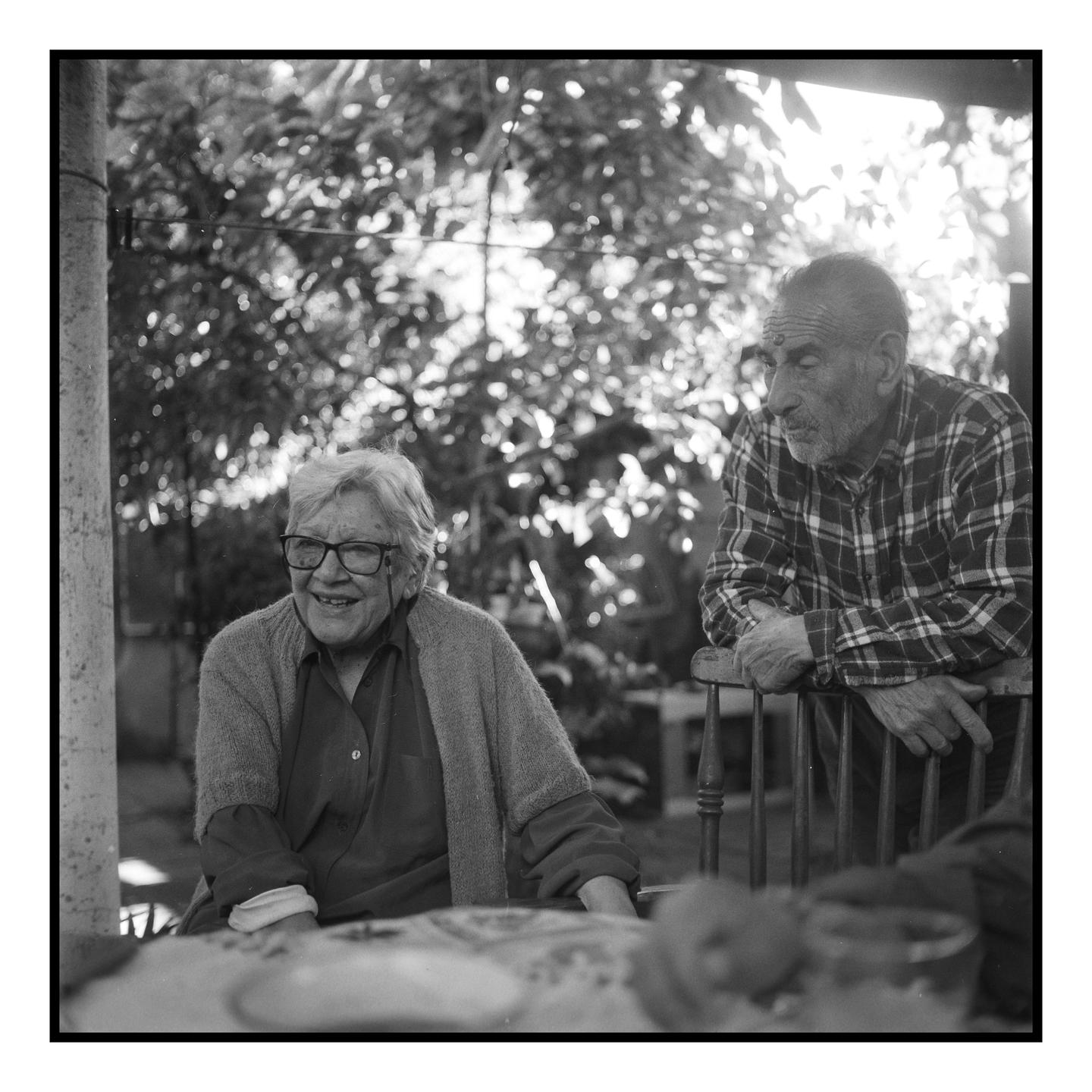 A woman in glasses smiles at a man leaning on a chair in a garden.