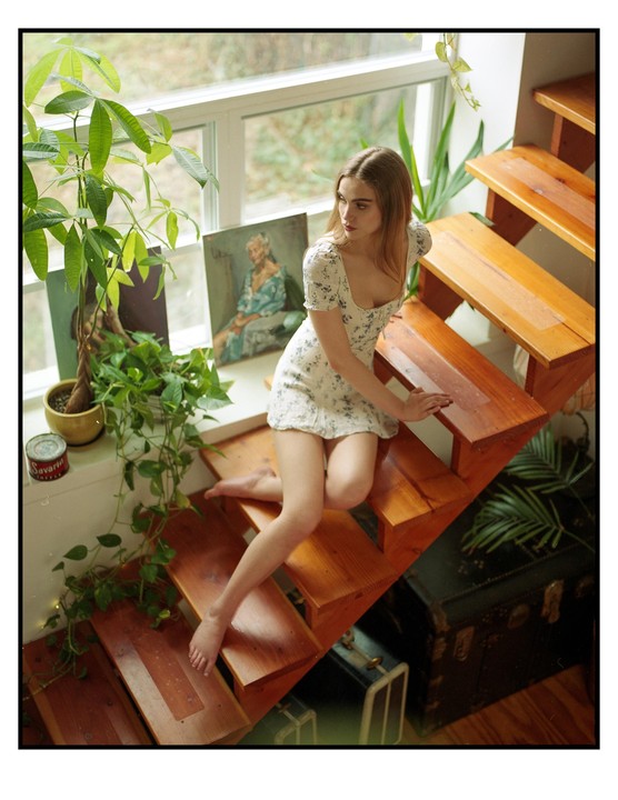 A woman in a white floral dress sits barefoot on wooden stairs next to a window with plants and paintings.