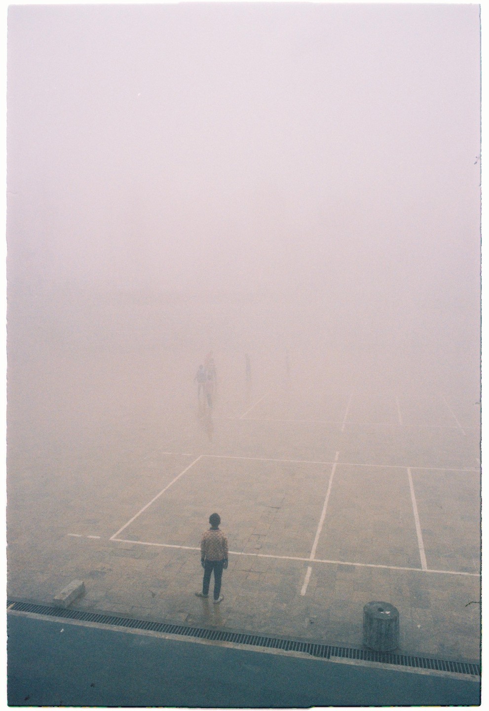 A person stands on a tennis court in thick fog, looking towards other blurry figures in the distance.