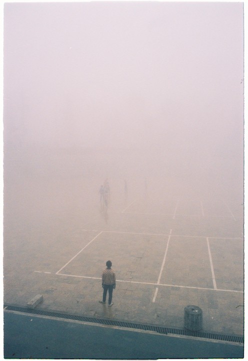 A person stands on a tennis court in thick fog, looking towards other blurry figures in the distance.