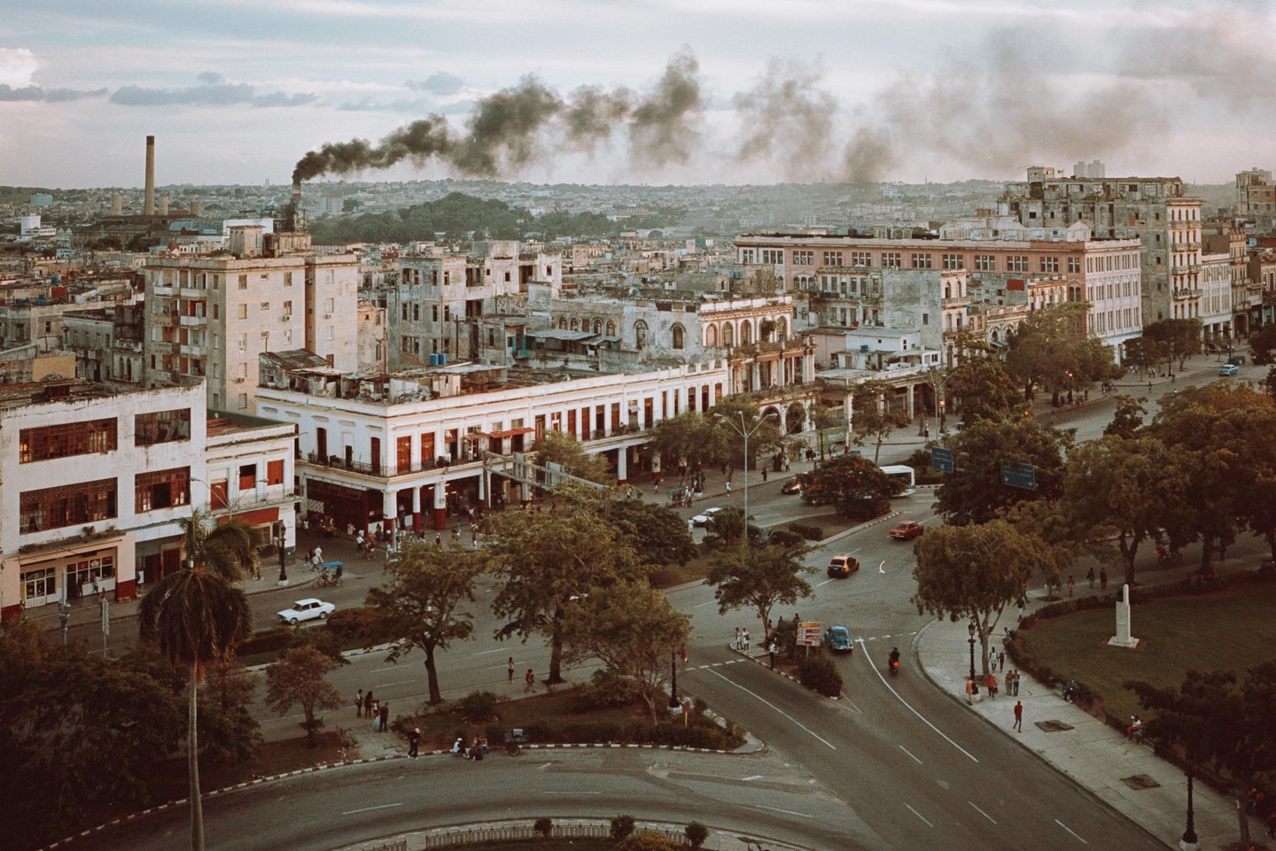 An aerial view shows a city with many buildings, trees, and cars on the street, with smoke rising from a chimney in the distance.