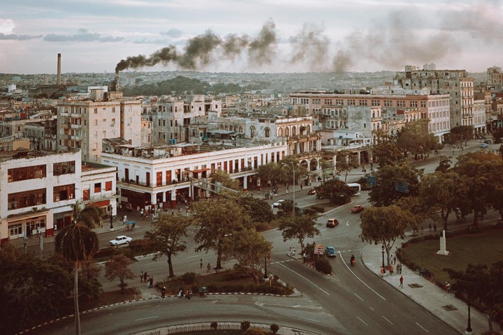 An aerial view shows a city with many buildings, trees, and cars on the street, with smoke rising from a chimney in the distance.