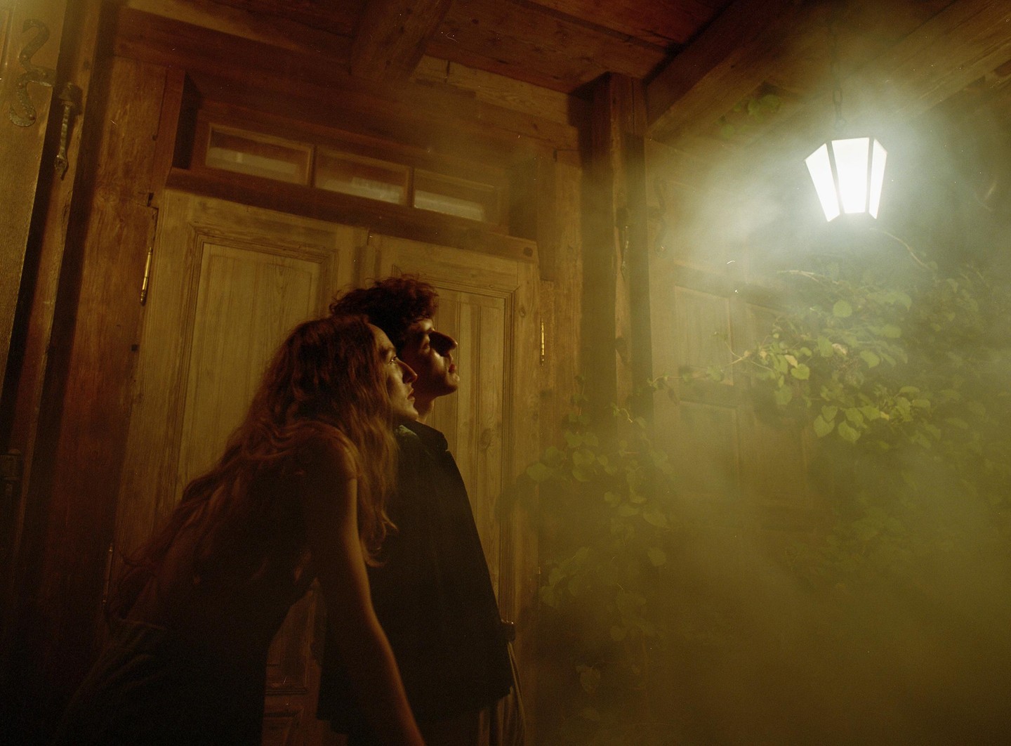 A couple looks up at a glowing lantern in a hazy, wood-paneled room with vines.