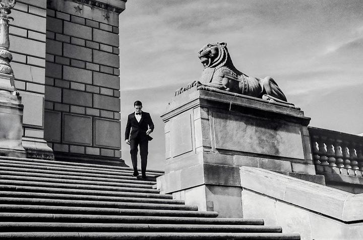 A man in a tuxedo descends a grand stone staircase next to a large stone lion statue.