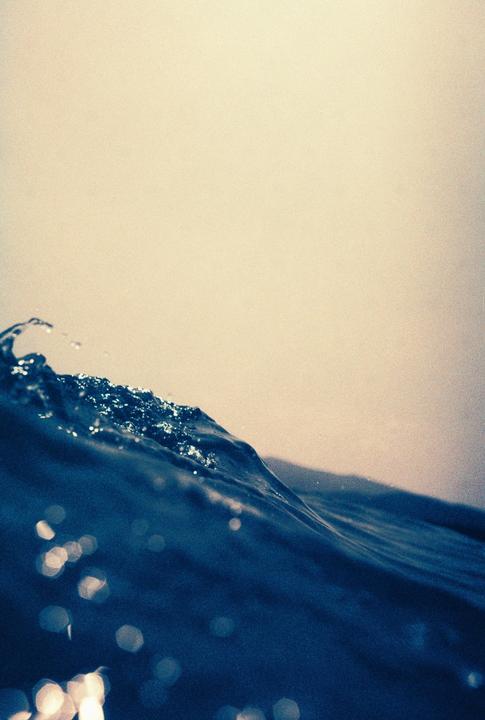 A close-up shot captures the crest of a dark blue ocean wave with sparkling water droplets against a light sky.