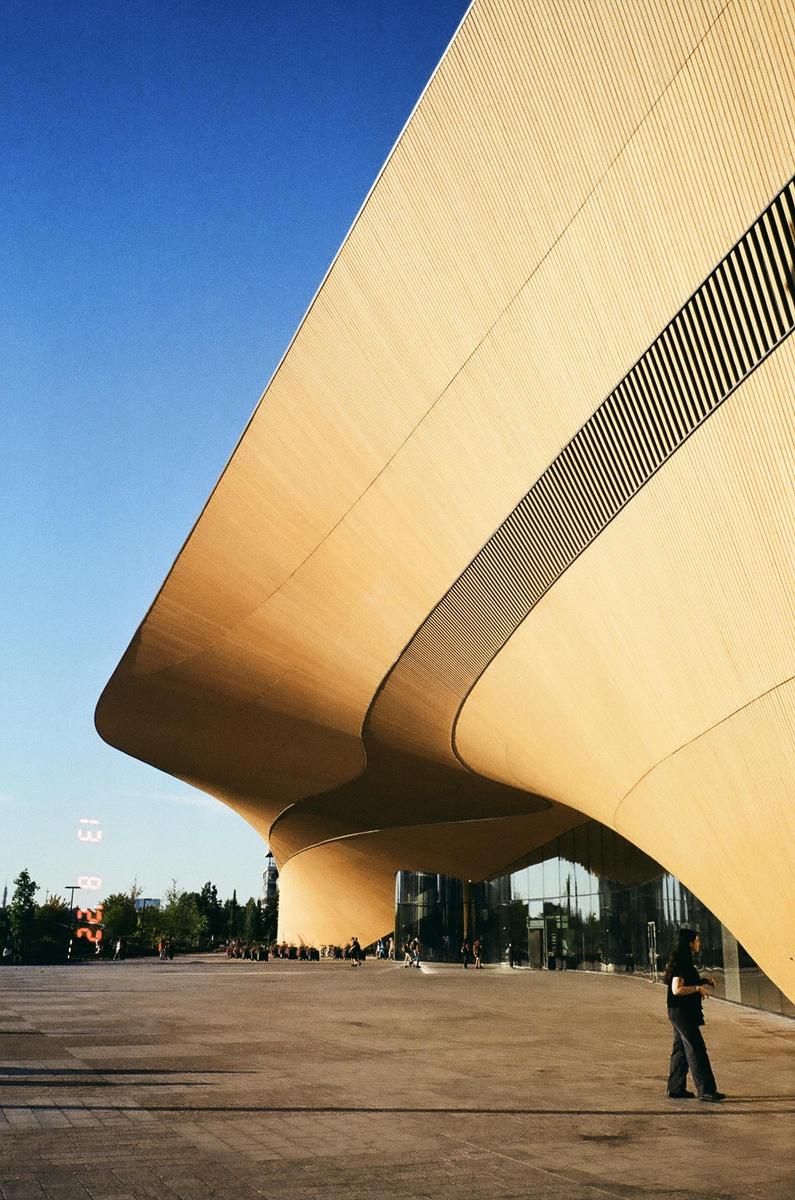 A person walks past a modern building with a curved wooden facade under a clear blue sky.
