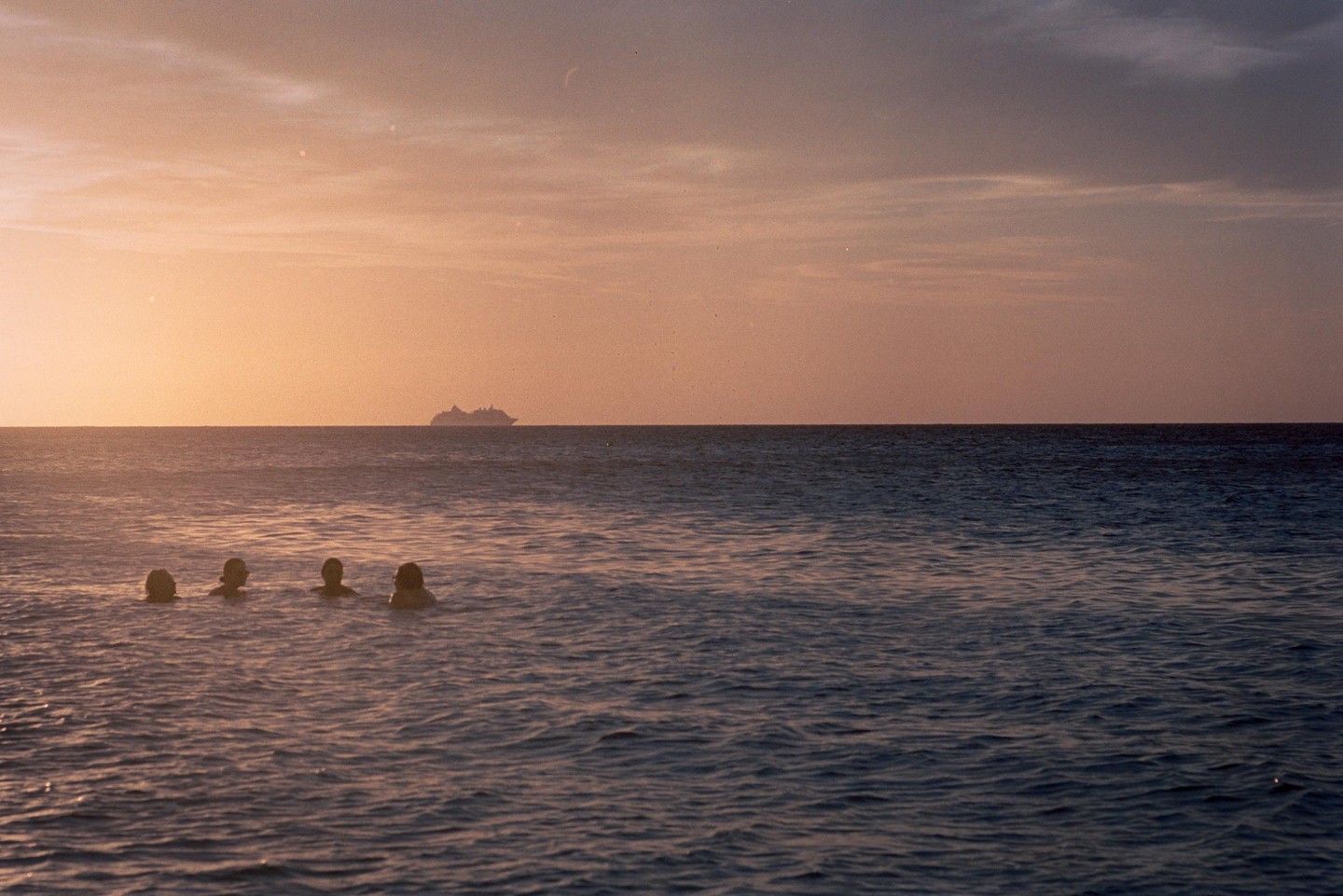 Four people swim in the ocean at sunset with a cruise ship on the horizon.