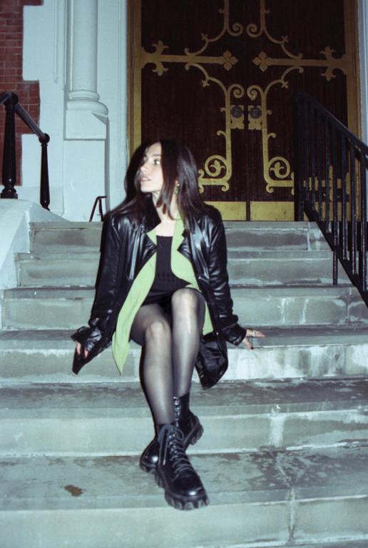 A woman wearing a black leather jacket and black boots sits on stone steps in front of a building with an ornate wooden door.