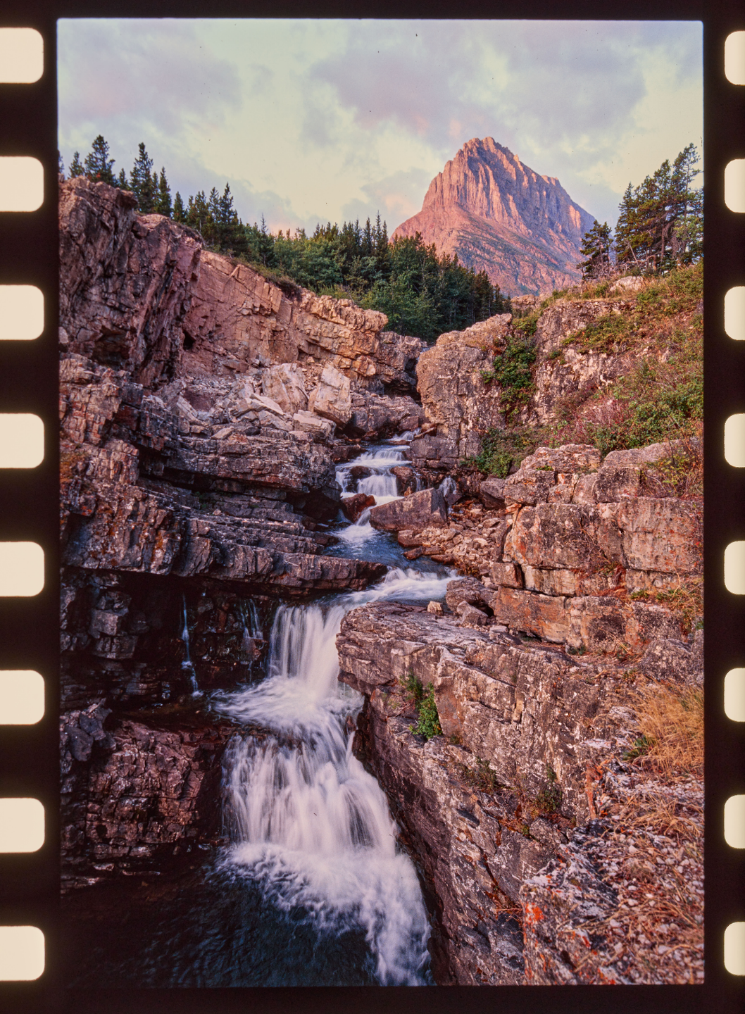 A waterfall cascades down a rocky gorge with a mountain and trees in the background.
