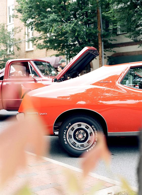 Two classic cars, one red and one orange, are parked on a street with their hoods open while people look at their engines.