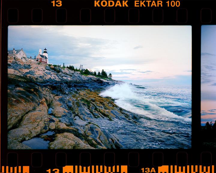 A lighthouse and buildings stand on a rocky coast as a wave crashes against the shore.