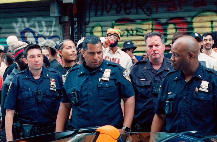 Several police officers stand in a crowd of people on a street with graffiti on a metal storefront gate.