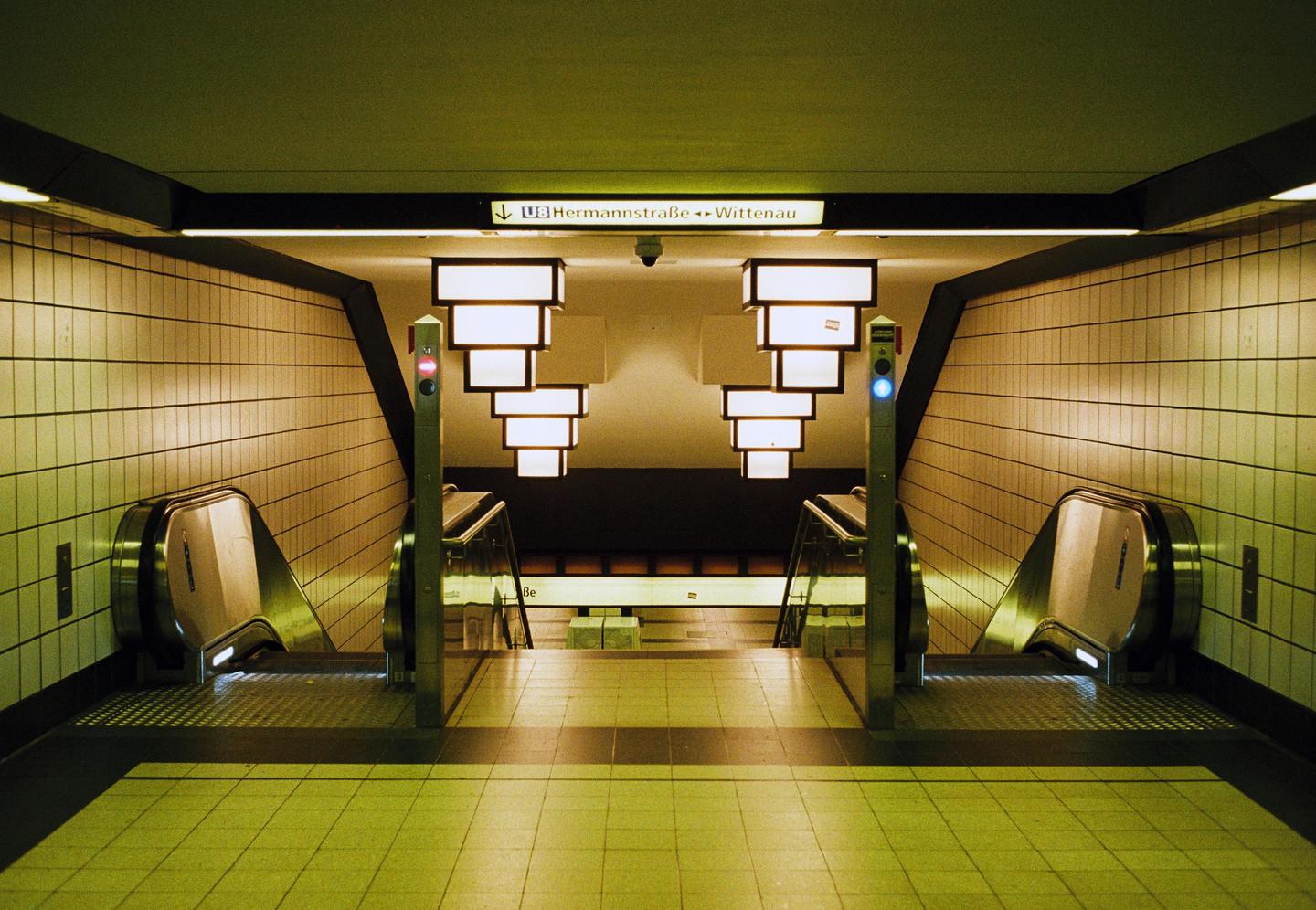 Two escalators descend into a subway station with tiled walls and illuminated signs.