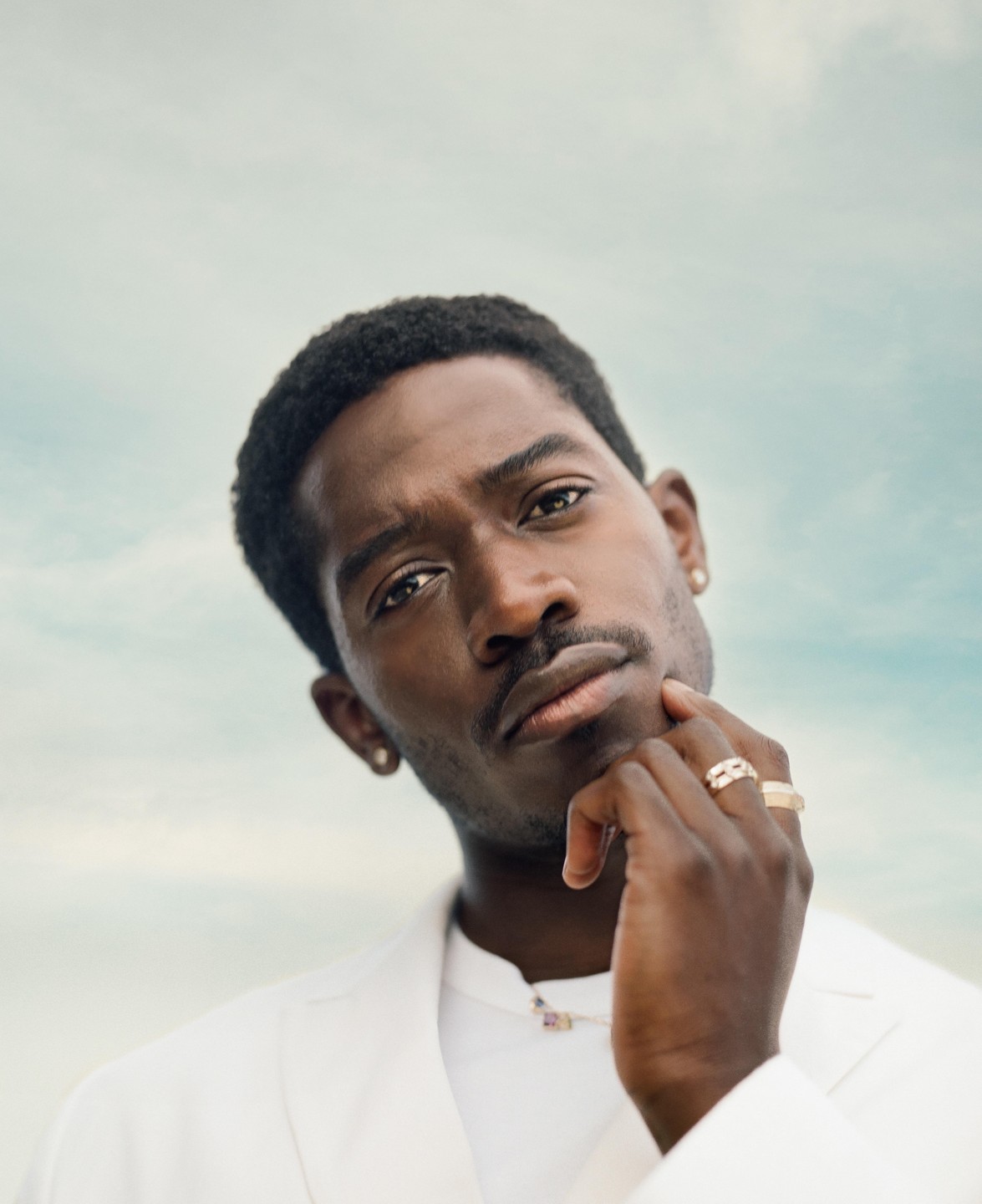 A man in a white jacket with rings on his fingers poses against a light blue sky.