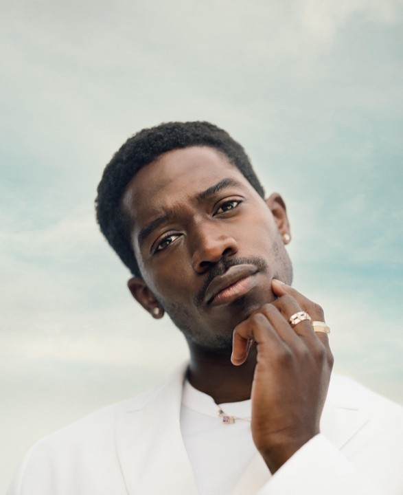 A man in a white jacket with rings on his fingers poses against a light blue sky.