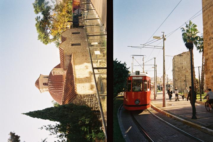 A red tram is on a track next to a stone building with a domed roof and a street with pedestrians.