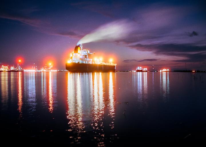 A large ship with bright lights and smoke coming from its chimney is on the water at night, with other lights visible in the distance and reflections on the water.