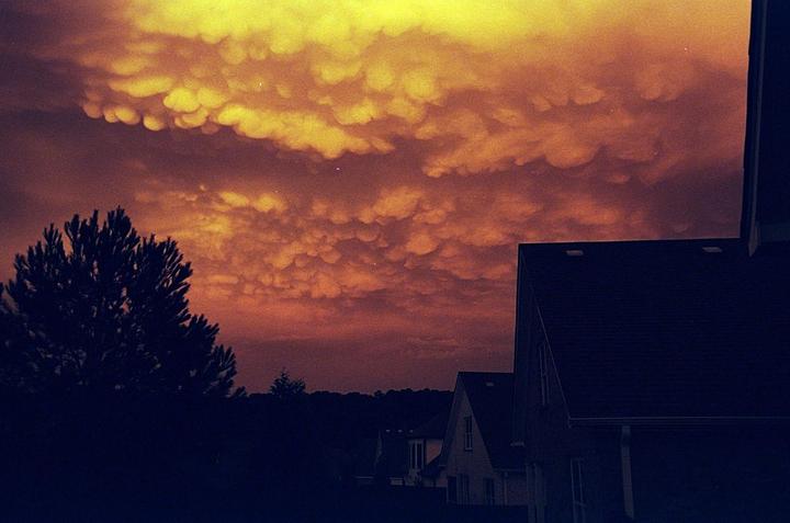 A dramatic sky with mammatus clouds hangs over silhouetted houses and trees at dusk.