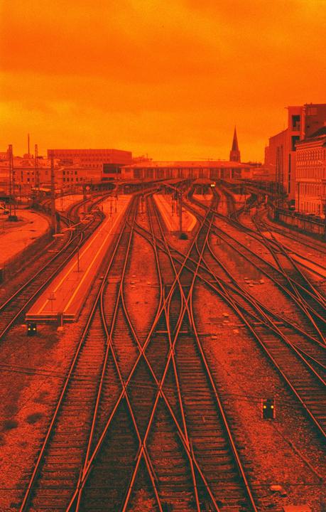 A train station with multiple tracks and platforms is visible under an orange sky.