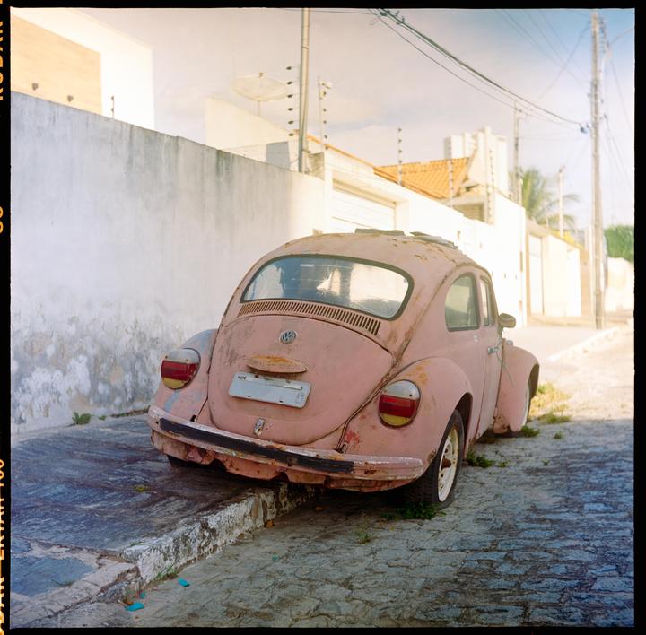 A pink, rusty car is parked on a cobblestone street next to a white wall.