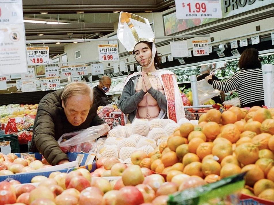 A man in a pope costume stands in a grocery store aisle next to a man selecting fruit.