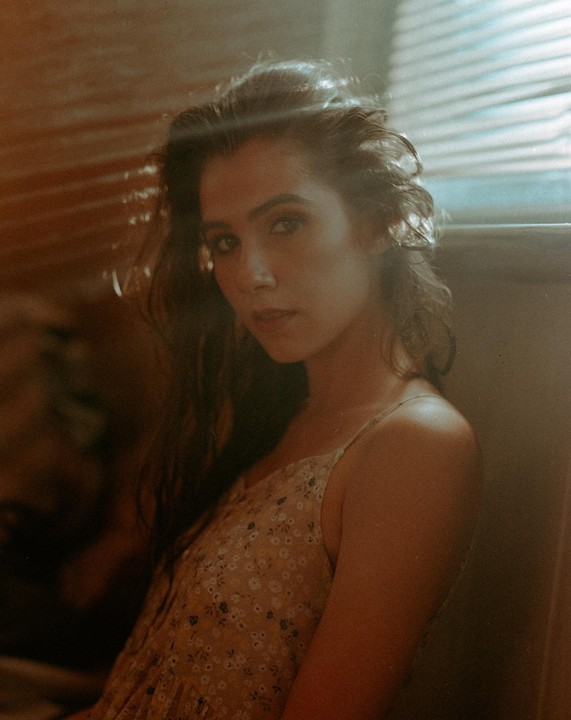 A woman with long, dark hair and a floral dress looks directly at the viewer, with light filtering through blinds in the background.