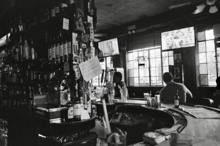 Two men sit at a bar with bottles of alcohol on shelves behind them and televisions above the windows.