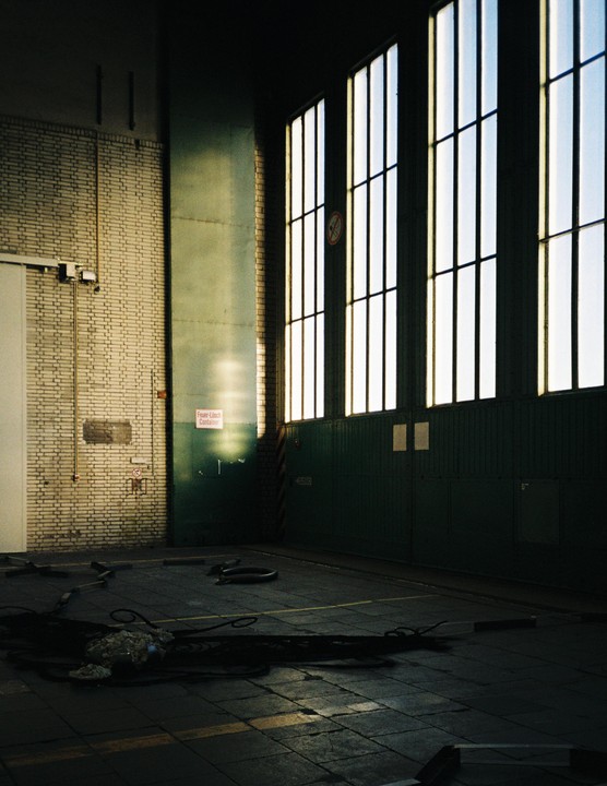 Sunlight streams through tall windows into a large, empty industrial building with a brick wall and a dark floor.