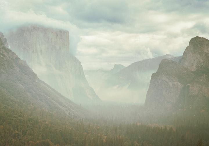 A hazy valley with a forest and mountains is visible under a cloudy sky.