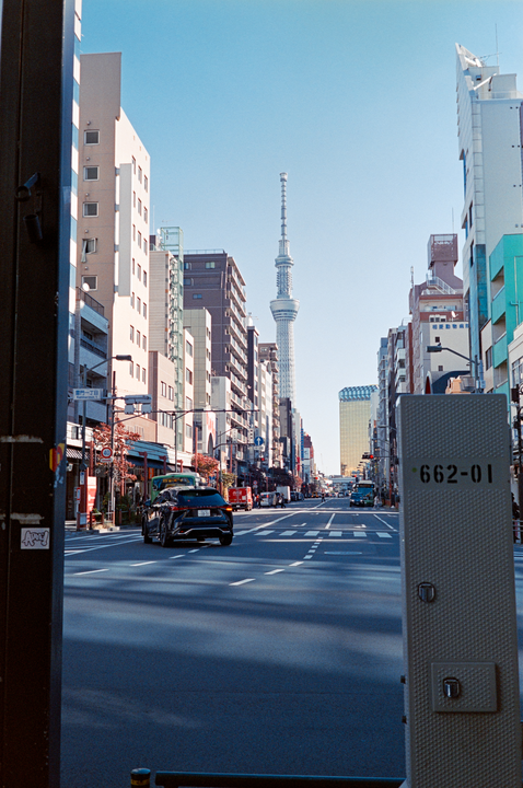 A street in a city is lined with buildings and cars, with a tall tower visible in the distance under a clear blue sky.