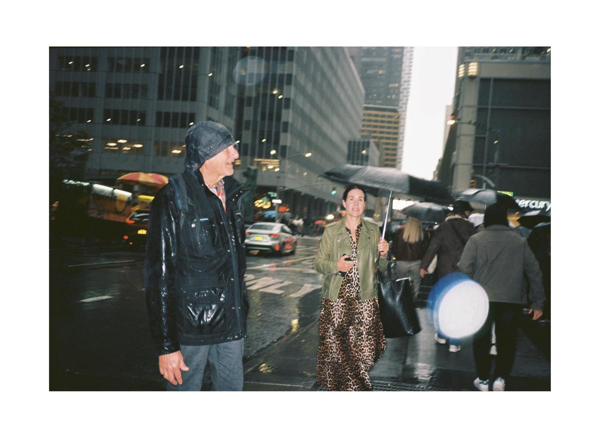 A man in a rain jacket and a woman with an umbrella walk on a city street in the rain.