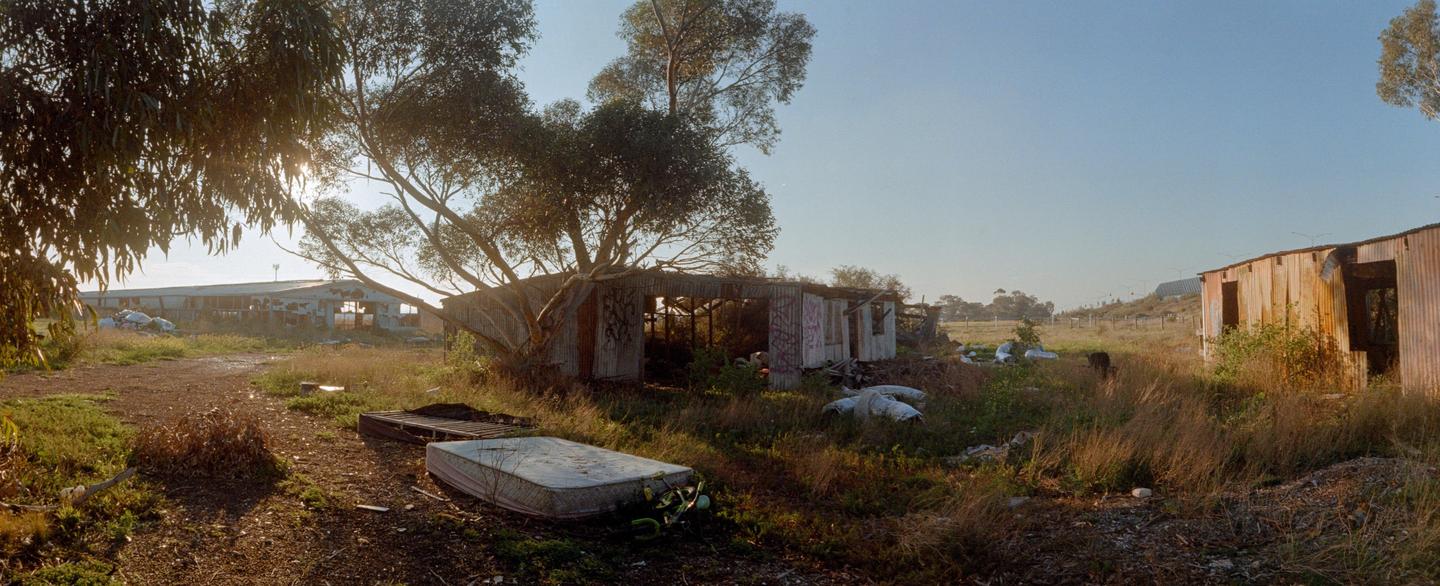 A panoramic view shows several dilapidated structures and an old mattress in a field with trees under a clear sky.