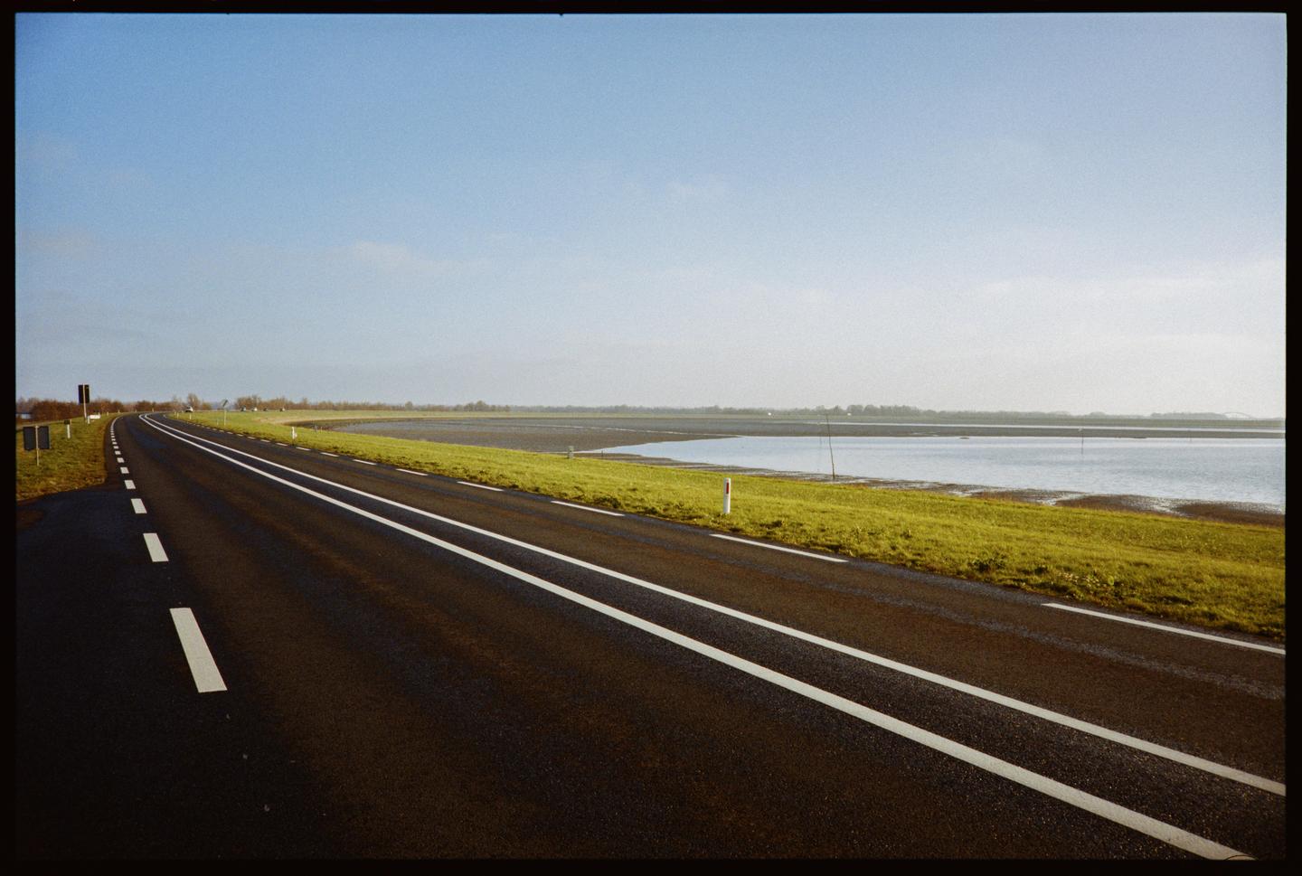 A road with white markings curves alongside a body of water with green grass on its bank under a clear sky.