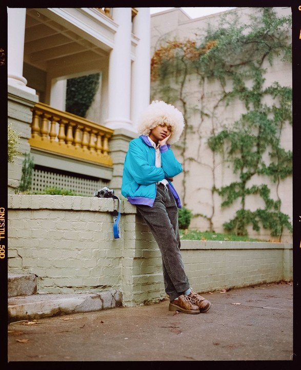 A person with a large afro hairstyle leans against a low wall next to a building with columns and a yellow railing.