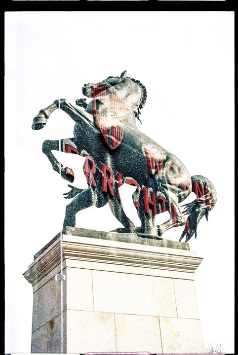 A double exposure shows a horse statue with red text overlaid on its body, standing on a stone pedestal.