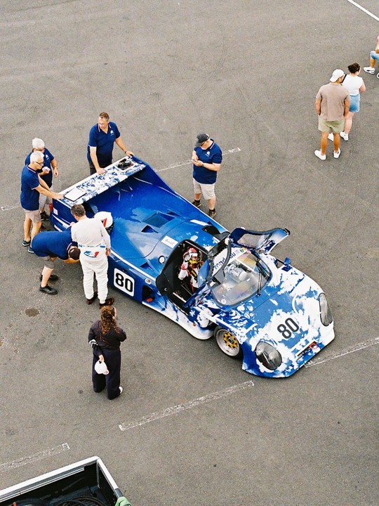 A blue and white race car with the number 80 is surrounded by several men in a pit area, with a woman standing nearby.