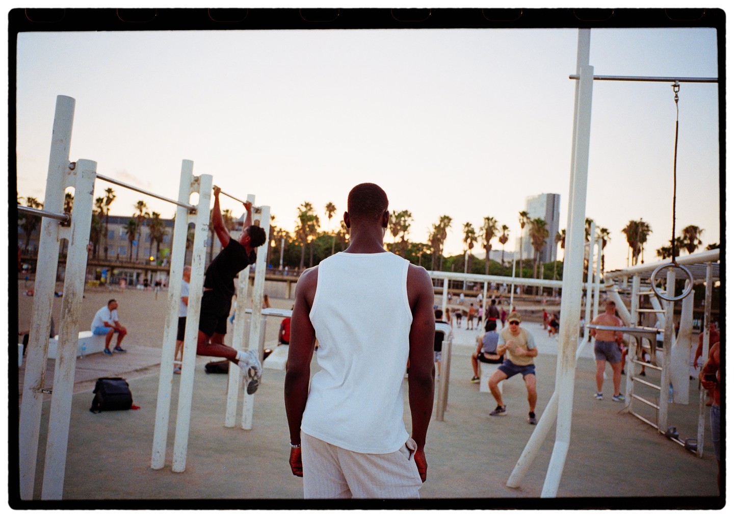 A man in a white tank top stands with his back to the camera, observing other men exercising on outdoor gym equipment at sunset.