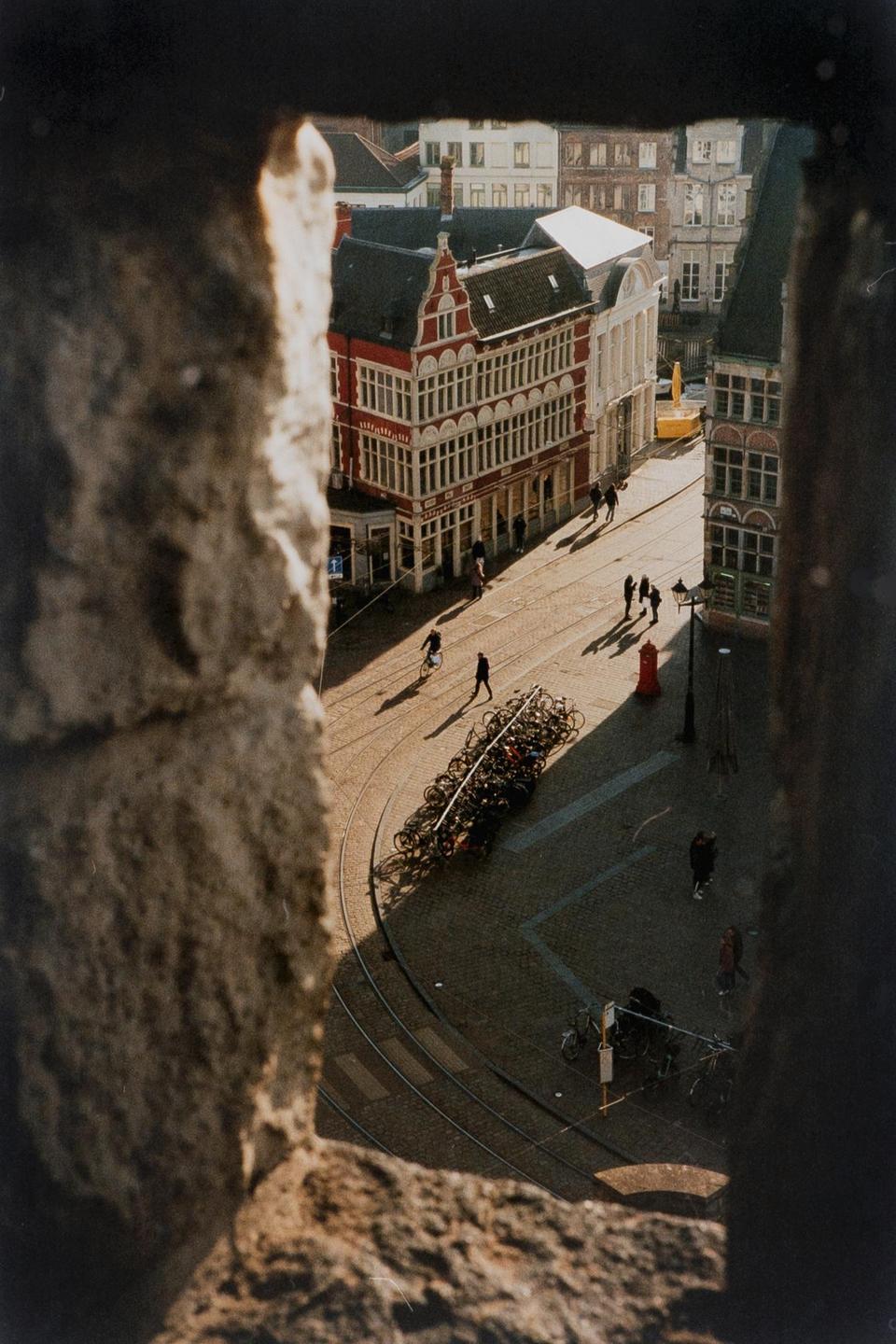 A high-angle view through a stone opening shows a street with buildings, people, and bicycles.