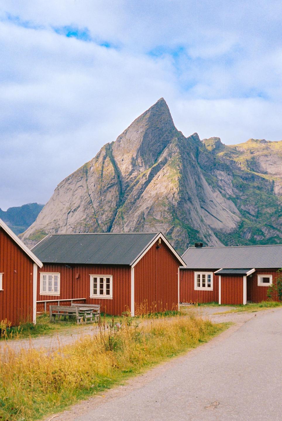 Red cabins with white trim line a dirt road with a large mountain in the background under a cloudy sky.