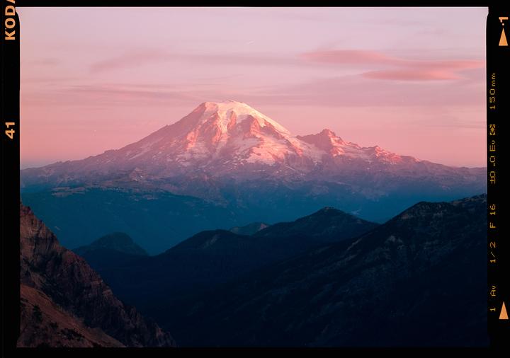 A snow-capped mountain is illuminated by the rising sun, casting a pink glow on the sky and clouds.