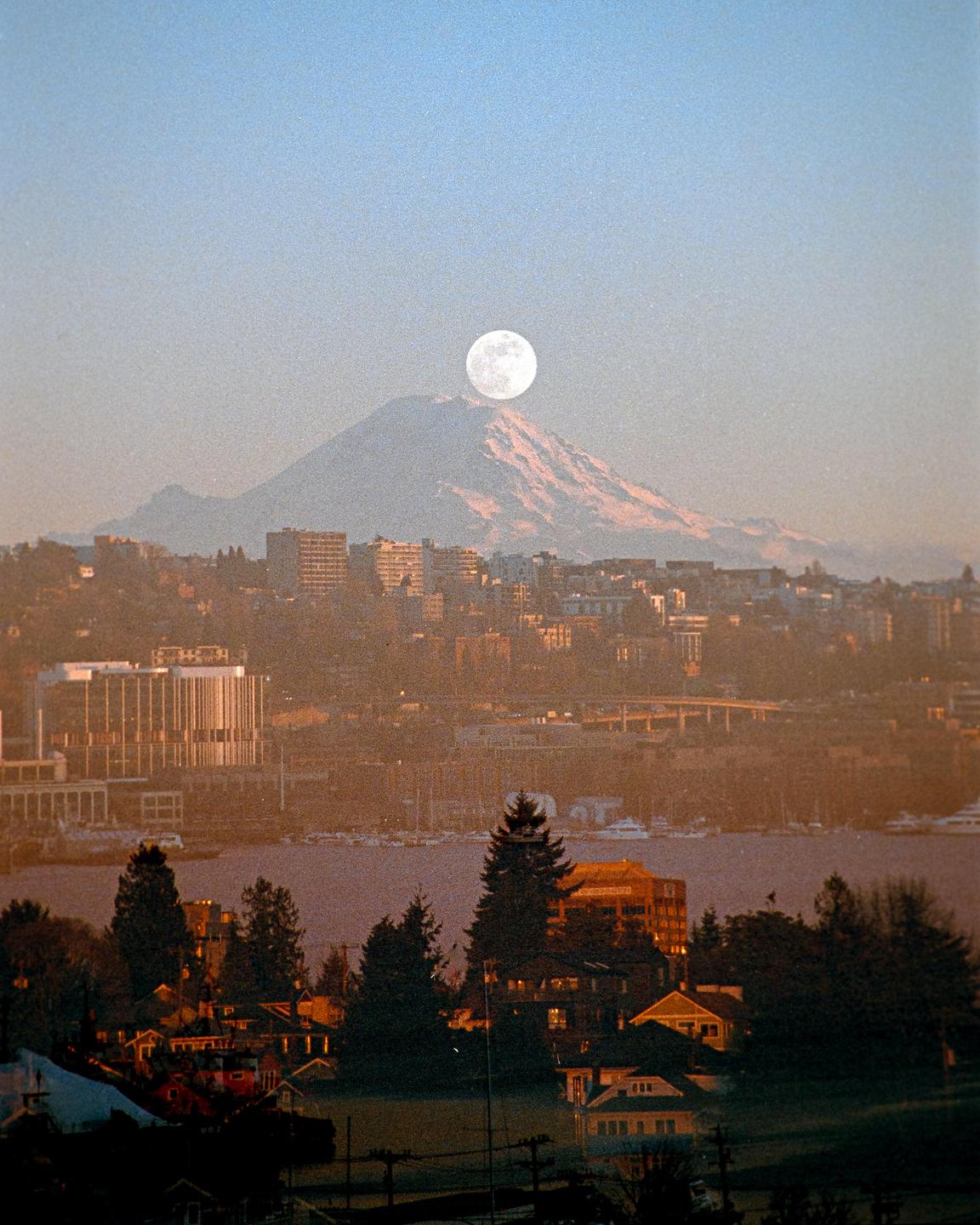A full moon rises over a snow-capped mountain behind a city and body of water.