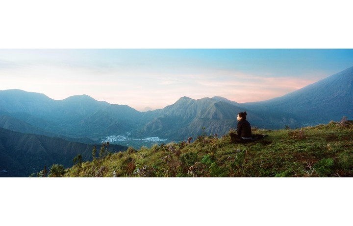 A woman sits on a grassy hill overlooking a mountain range and a small town at sunrise.