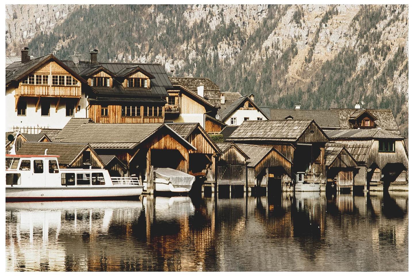 A village with wooden houses and boat sheds is reflected in the water of a lake, with a mountain in the background.